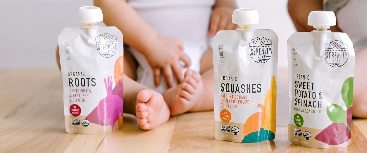 Spouted baby food pouches standing on a wooden floor beside a seated child